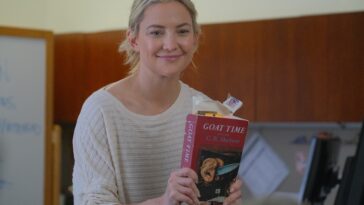 Kate Hudson as Prof. Cleary holds up a copy of the novel Goat Time.