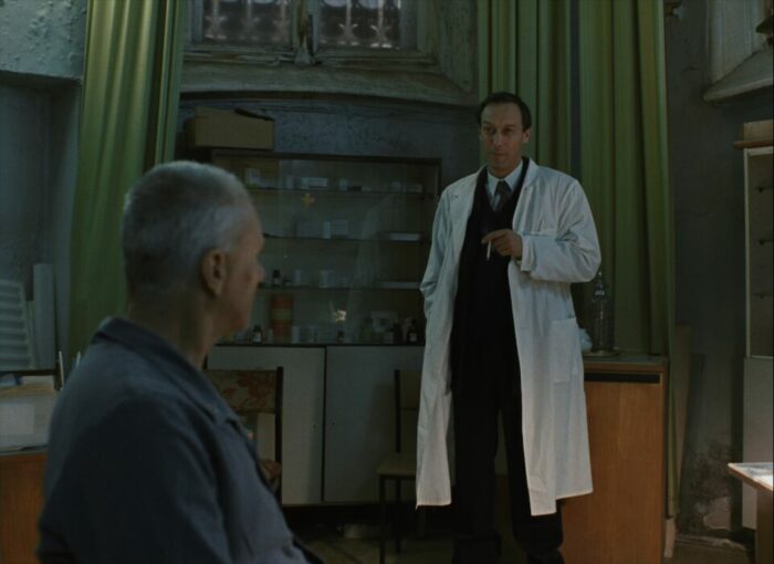 A psychiatrist in a white lab coat (Oleg Yankovskiy as Smirnov) speaks to a seated patient (Malcolm McDowell as Timofeyev) in his office.