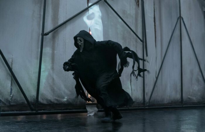Ghostface runs after someone with a large screen in the back, protecting a movie of Ghostface.
