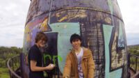 two teenagers looking at the camera on the top of a graffiti covered watertower, one of them is holding the camera pointed at themselves.