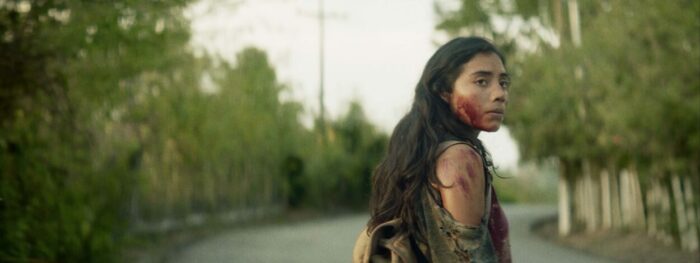 A bloodied woman looks back at the camera as she walks along an empty road. 