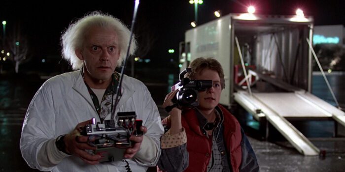 Doc with a remote control and Marty with a videocamera watching the DeLorean in a parking lot