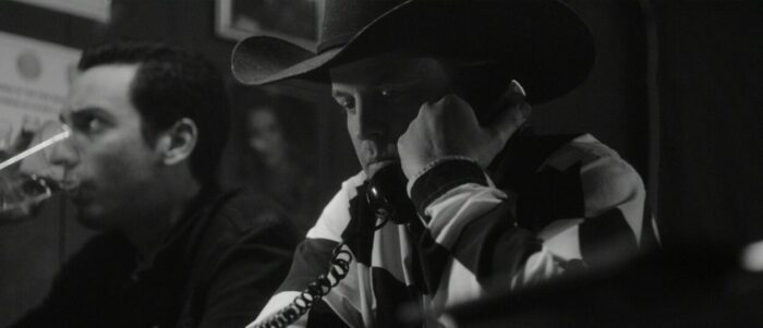 Mickey Reece as Troyal Brooks, wearing a cowboy hat and talking on a telephone in a bar.