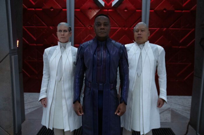 A purple scientist is flanked by two aides in white tunics.