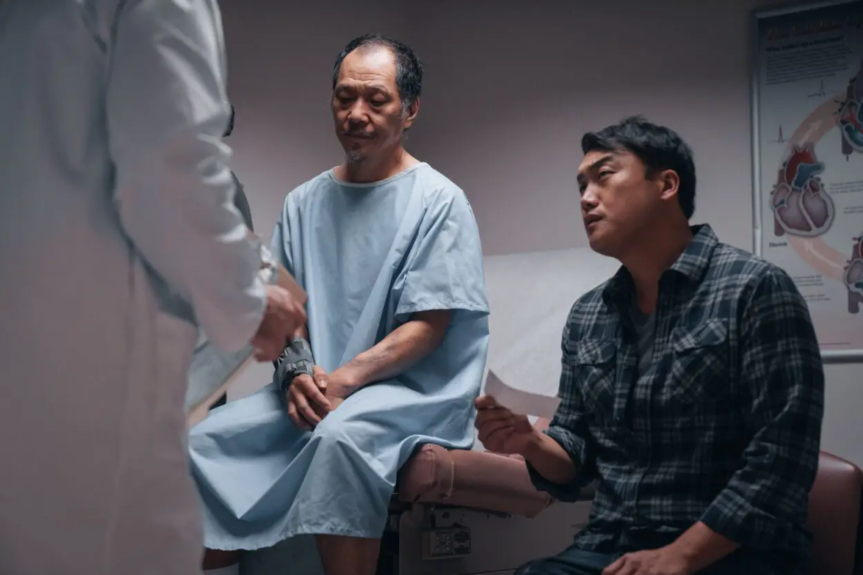 Cher (Perry Yung), wearing a hospital gown, sits in the doctor's office with his son Thai (Doua Moua) as they receive news of Cher's ailing health.