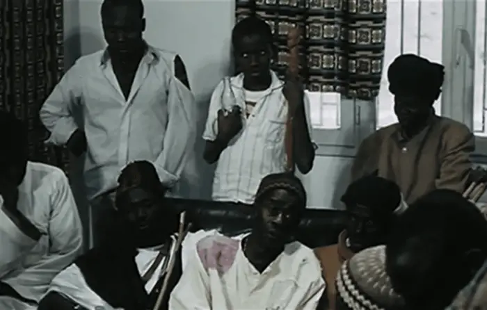 Screenshot from Xala showing a group of men, with some sitting on a couch in the foreground and some standing up behind them. 