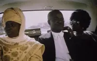 An African man in a tuxedo rides in the back seat of a limousine between two women.