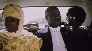 An African man in a tuxedo rides in the back seat of a limousine between two women.