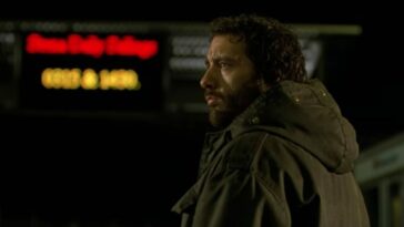 Will, in a coat with a beard, in front of a neon sign looks to his left.