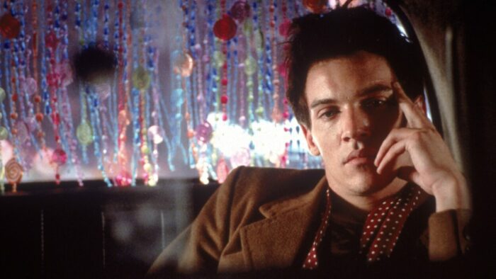 Davey sits in the back of a cab with his left hand against his face. The back of the cab has beads hanging from the ceiling.