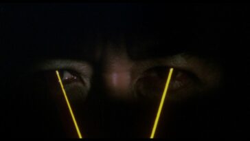 Close-up on Carlos in shadow with yellow lasers emitting from his eyes.