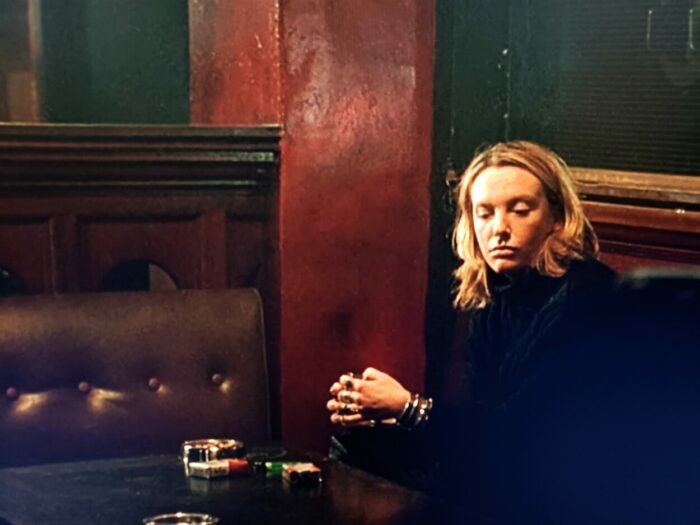 Toni Collette as Mandy Slade, looking depressed, dressed all in black in a dimly lit dive bar, in Velvet Goldmine (1998)
