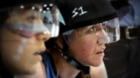 Two women in roller derby helmets stare off to camera right.