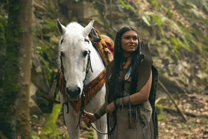 A Native American woman smiles and stands next to her horse.