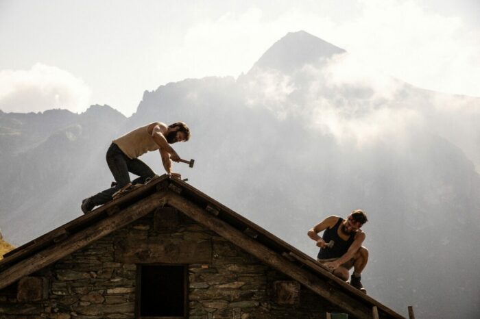 Two men work on the roof of a cabin in the mountains.