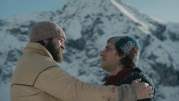 Two men in beards, hats, and coats embrace on a mountain.