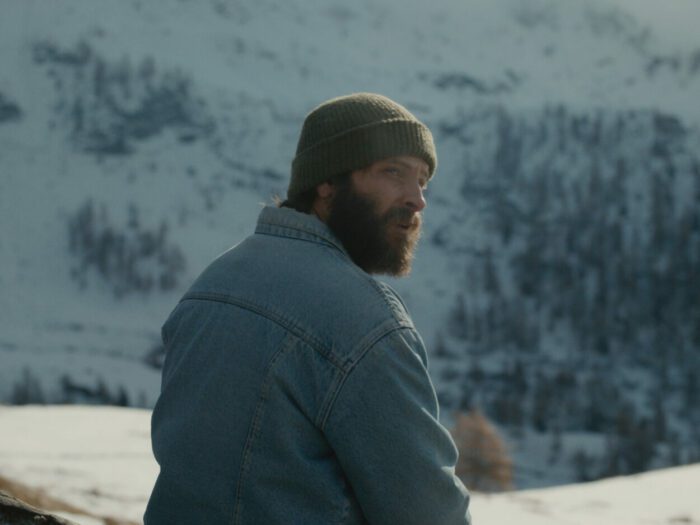 A bearded man wearing a cap and jacket in front of a snowy mountain.