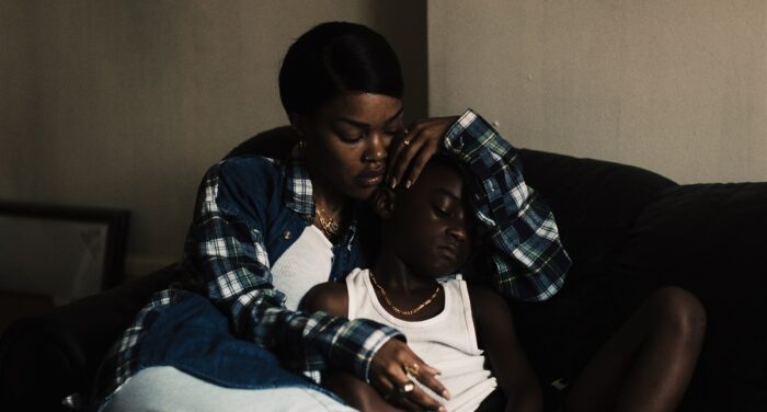 Inez and Terry (played by Aaron Kingsley Adetola) cuddling on a black couch in a dimly lit room. Inez has one hand draped over Terry's head while the boy looks down at his hands.