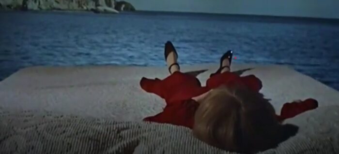 film scene from rosemary's baby, depictng a woman in a red dress (or perhaps bathed in blood) on a bed at the edge of an open sea