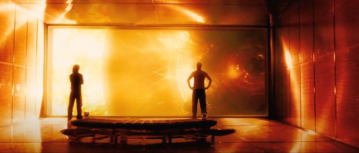 Two men look out a large window at a spaceship, the room glows as the sun shines into it bathing it in red, yellow and black 