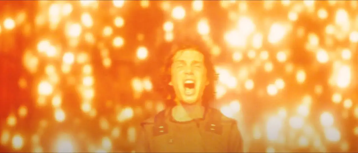 A man stands and screams as he is surrounded by light and glowing orbs, his hair is blowing back and his mouth is wide open 