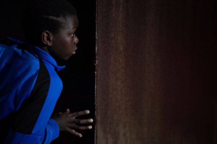 A young boy in the dark peers through a doorway.