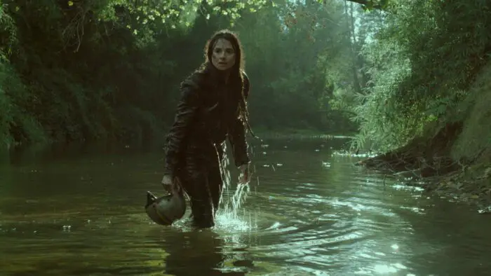 A woman wearing a motorcycle jacket and carrying a helmet emerges from a river.