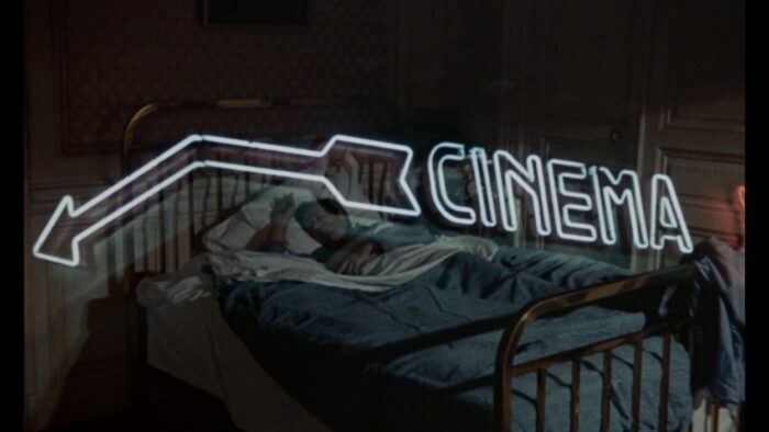 A man sleeps in a hotel bed as a neon cinema sign flashes over him.