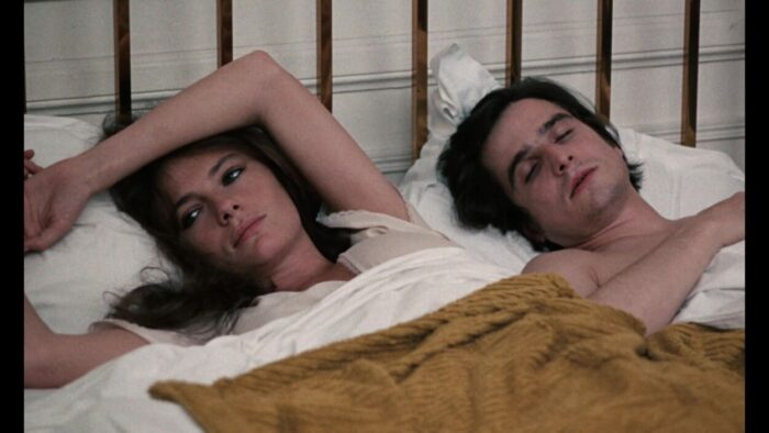 A man and a woman together in bed; the woman's eyes are open, the man's closed in sleep.