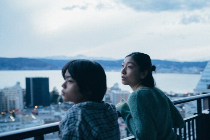 A woman and a child look out from a balcony over the skyline.