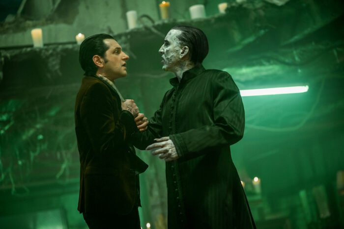 Teddy Lobo (Ben Schwartz), a white man with dark, slicked-back hair and a brown velvet blazer, grabbing the arm of Dracula, who is wearing a long black night coat and looks considerably more withered in the face, cheeks hollow and face adorned with dark holes. Dracula's arm is resting on Teddy's right shoulder. They are in Dracula's lair, lit in a ghostly green.