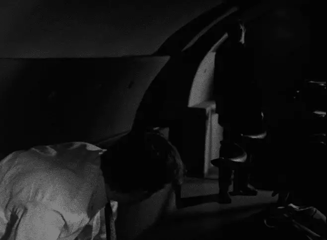 Screenshot of the movie Following showing a silhouette of a man in a dark room in the background and a man bending down a few feet away from him in the foreground. 