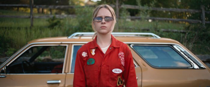 A woman in coveralls with badges stands with sunglasses.