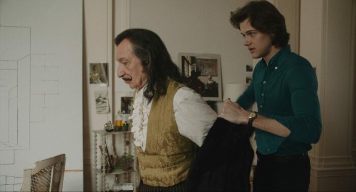 Medium shot of James (Christopher Briney) helping Dalí (Ben Kingsley) put on his coat in his art studio
