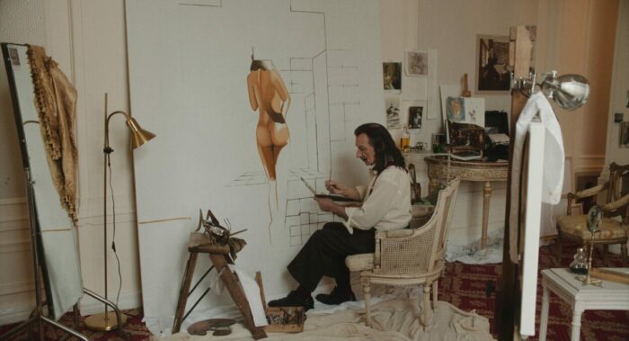Salvador Dalí (Ben Kinsley) sitting in an arm chair adding to a large painting of the back of a naked woman in his cluttered art studio. 
