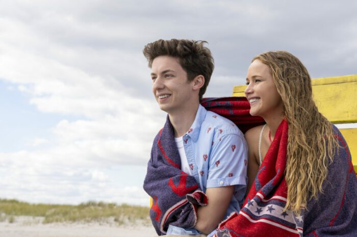 Percy and Maddie sit at the beach, covered with a blue and red towel that they are sharing as they smile.