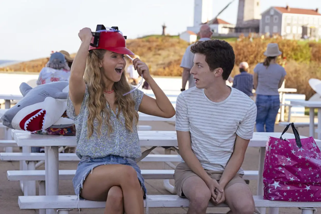 Maddie (Jennifer Lawrence) and Percy (Andrew Barth Feldman) sit on a bench outside while Maddie puts on a red novelty hat with slots to hold a drink with straws.