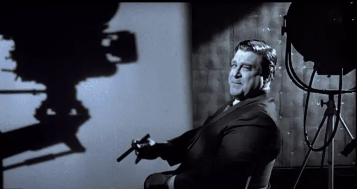 A filmmaker holding a cigar sits in a director's chair next to the silhouette of a movie camera