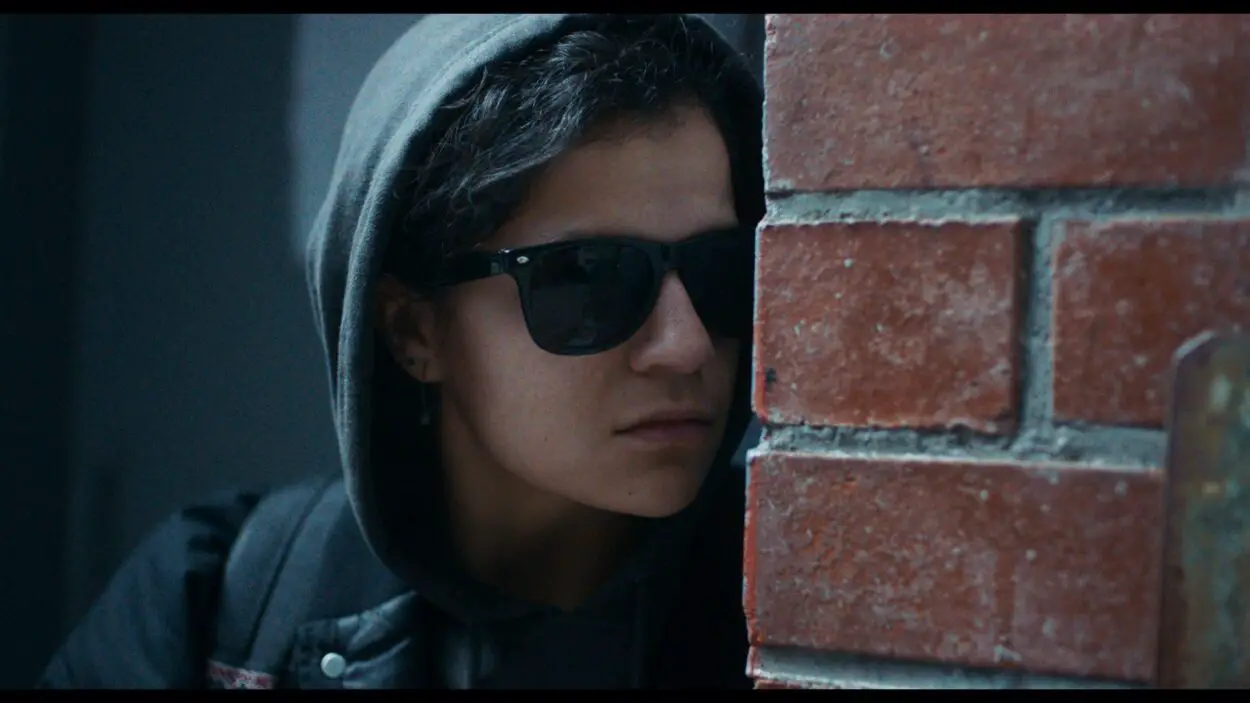 A woman wearing sunglasses peeks around a brick corner