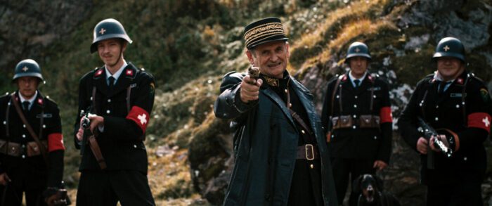 Still from the film MAD HEIDI showing five soldiers standing in a row. the Man in the middle, Commandant Knorr, points a gun forward toward the camera.