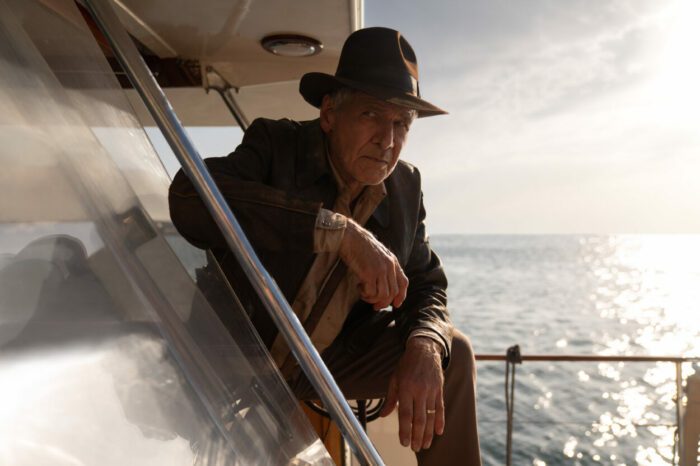 A man in a fedora and jacket leans outside a boat cabin door.