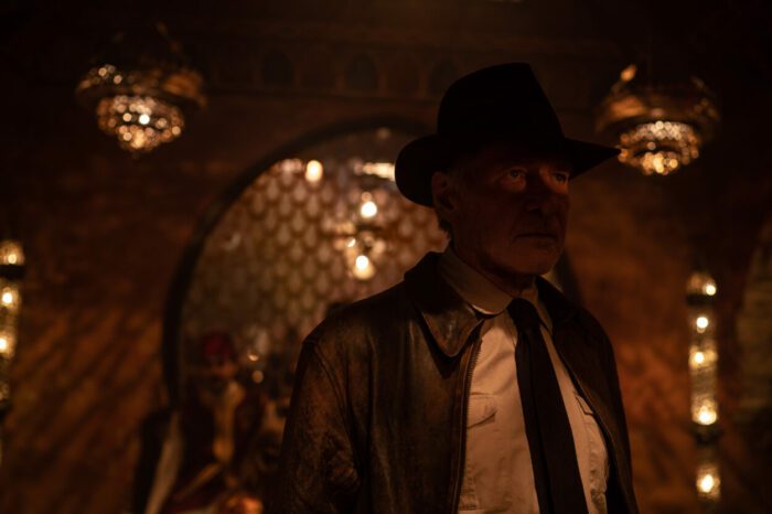 A man in a jacket and fedora stands in a room of shadows and entries.