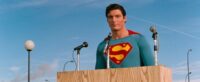 Superman stands at a podium of microphones