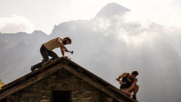 Two shirtless, bearded men work on a rooftop in the mountains.