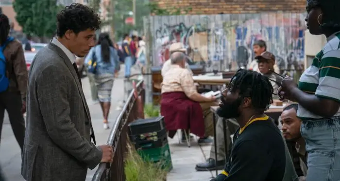Noah (Anthony Ramos) on the left, wearing a gray blazer, looking down at Reek (Tobe Nwigwe) from behind some railing outside of a restaurant's outside seating.