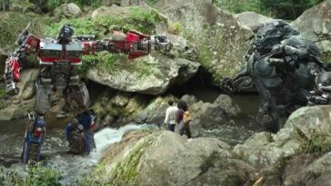 Optimus Prime (left) in the center of a rocky jungle stream, aiming his gun at Optimus Primal (right) while Noah and Elena (Dominique Fishback) watch from the center.