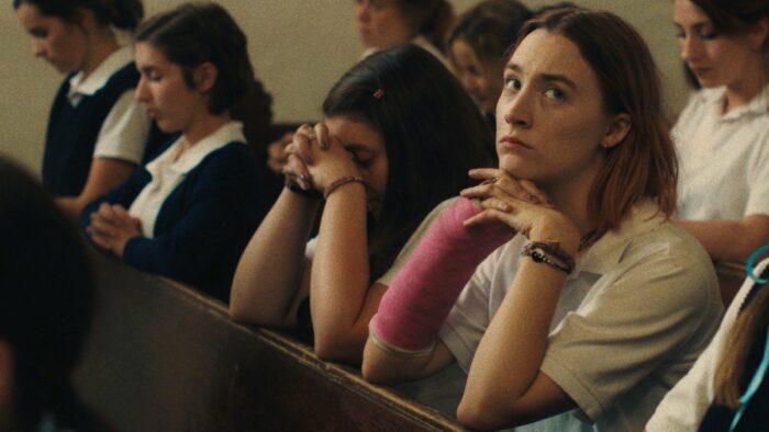 Lady Bird (Saoirse Ronan) with a pink cast in her arm kneeling at a pew looking up. 
