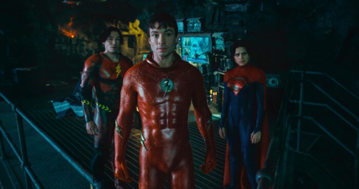 Two versions of Barry Allen, one wearing a in-universe accurate superset and another wearing a makeshift Batman suit, and Supergirl in the Batcave.