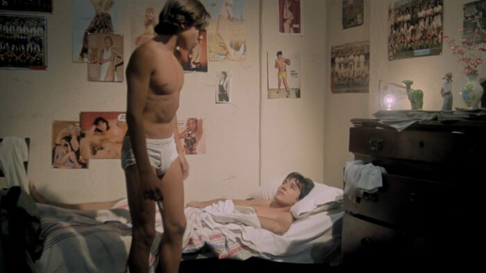 A teenage boy in his underwear stands next to another young boy in a bed.