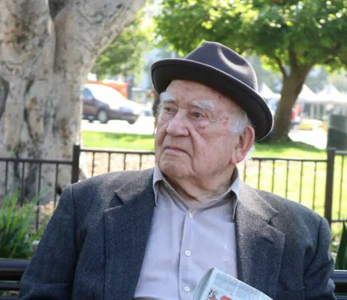 A senior gentlemen with a hat and jacket sits on a bench.